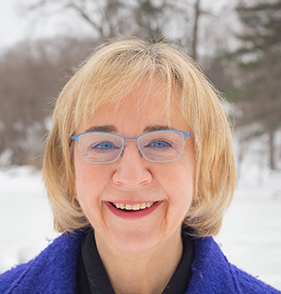 A headshot of Alice Diamond. She has short blonde hair and glasses, is wearing a dark blue coat and there is snow in the background.