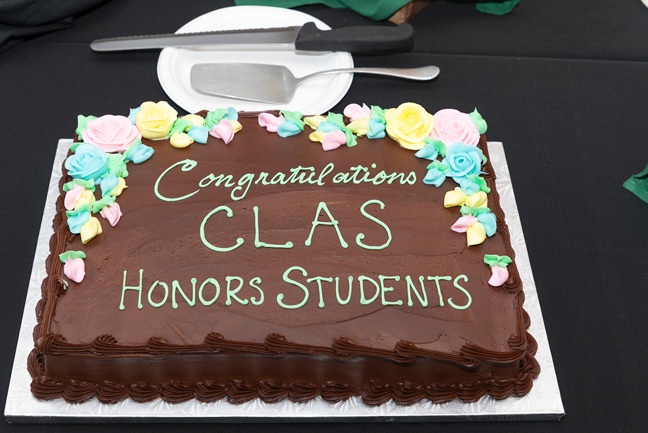 A photo of the cake from the 2022 Anual Honors Symposium. The cake is covered in brown frosting and says "Congratulations CLAS Honors Students" in green frosting. 