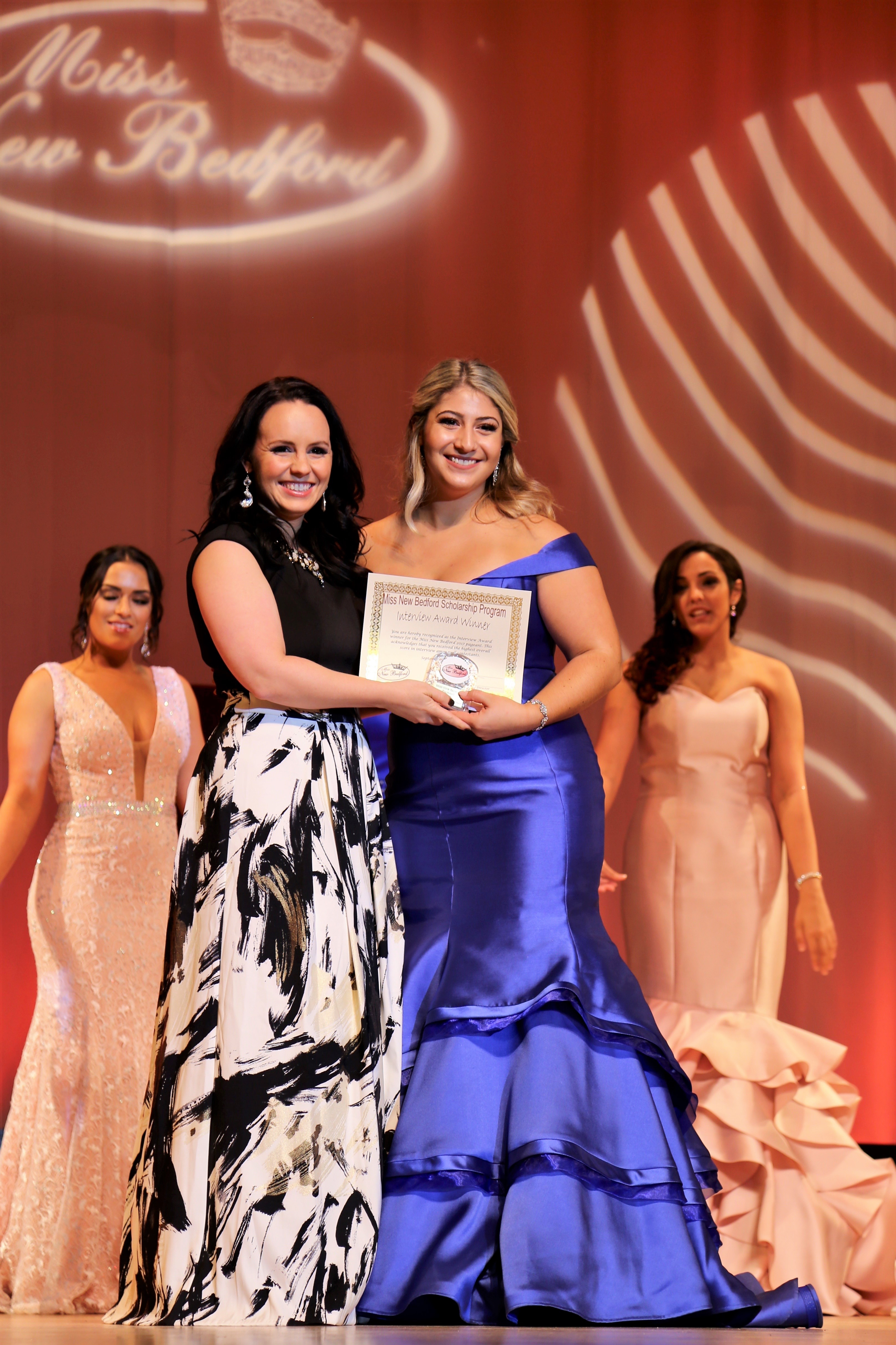 Kenzie Moniz receives the Interview Award standing on stage in a floor-length shiny purple dress.