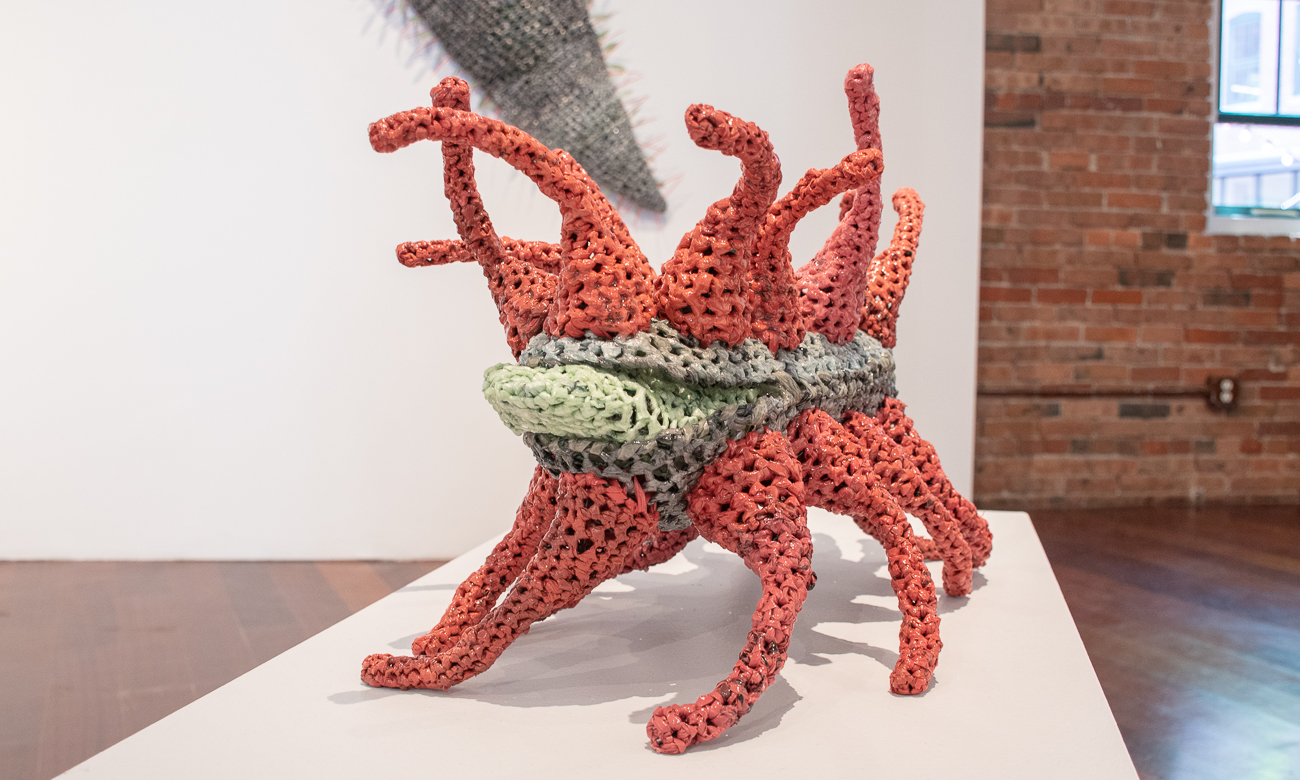 Artwork titled Germinate, made from red and green crocheted plastic. Appears to be standing on multiple legs with other tenticals come out of the top.