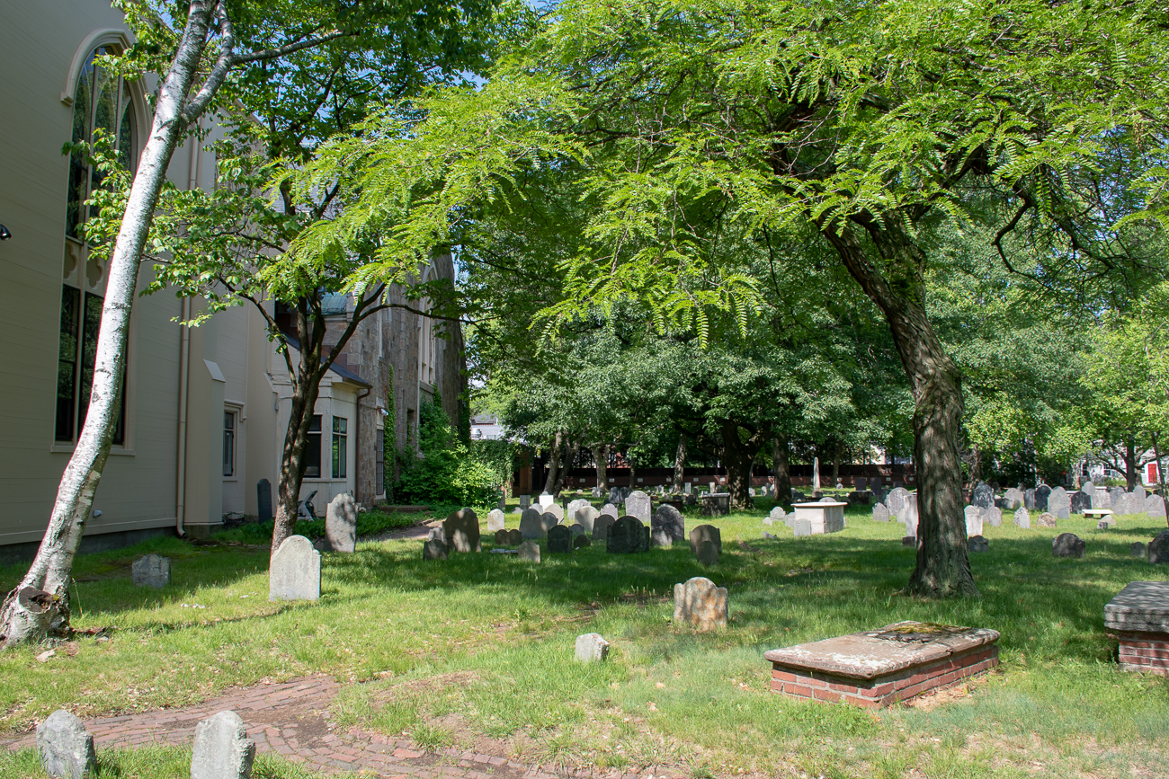 A view of graves and the side of the church next to the cemetery, with a lush tree casting shade.