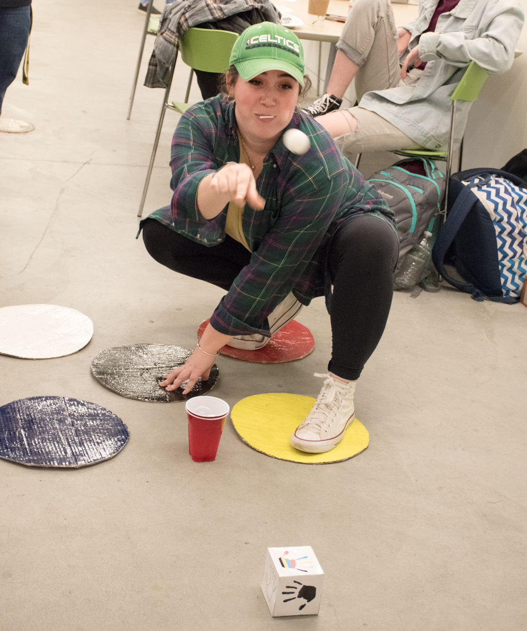 A girl crouches down on several colored Twister cirlces and throws a ball.