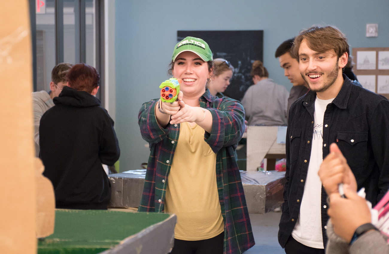 A student points a toy gun at a game while another male student looks on smiling.