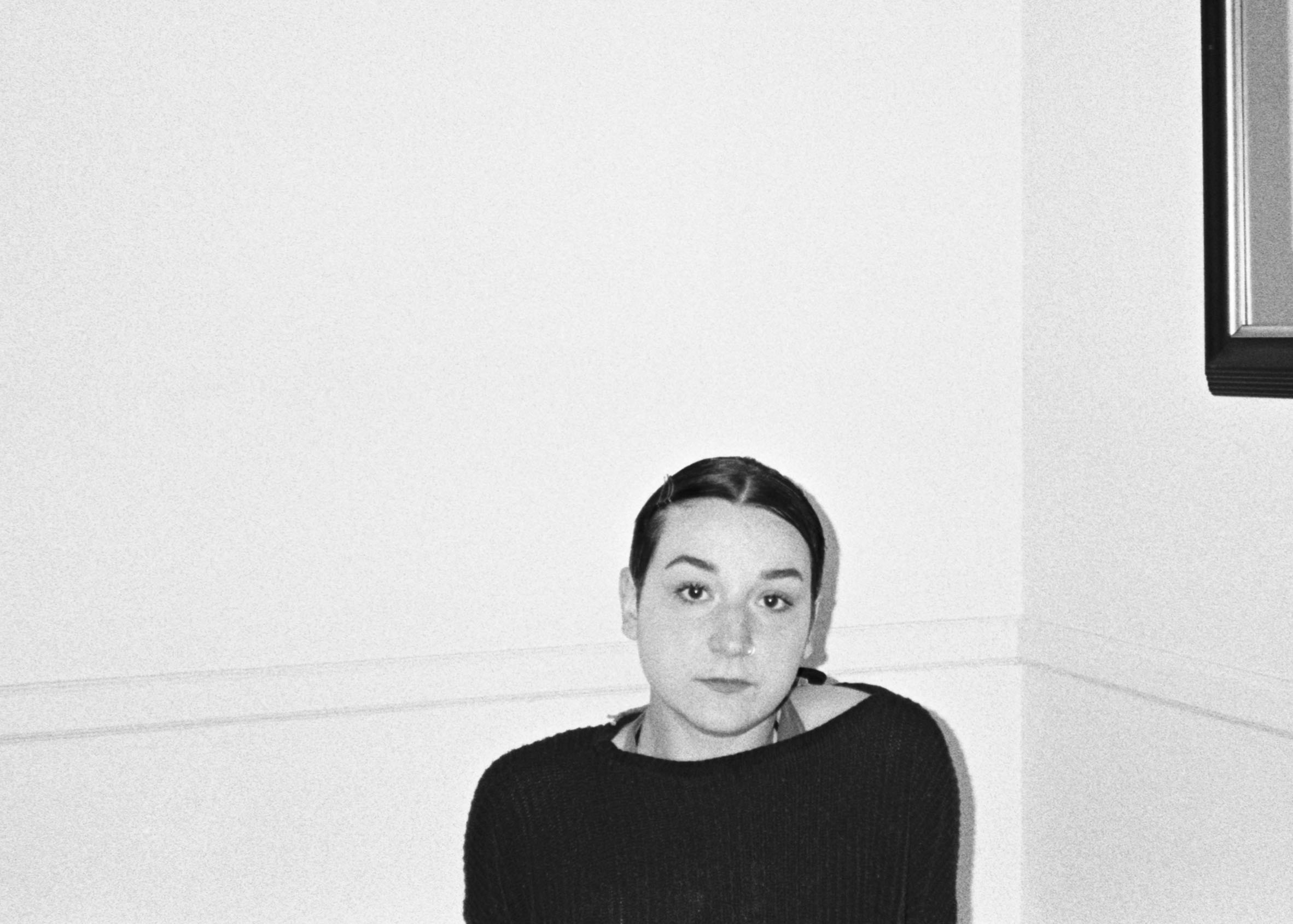 black and white photo of young woman sitting in empty white room wearing a black sweatshirt