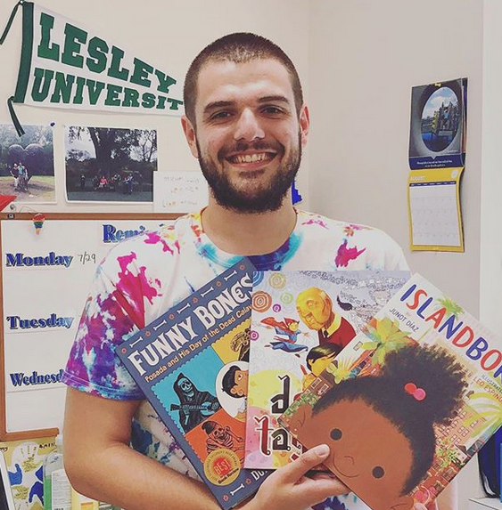 Man in a tie dye t-shirt holding up three books in a classroom