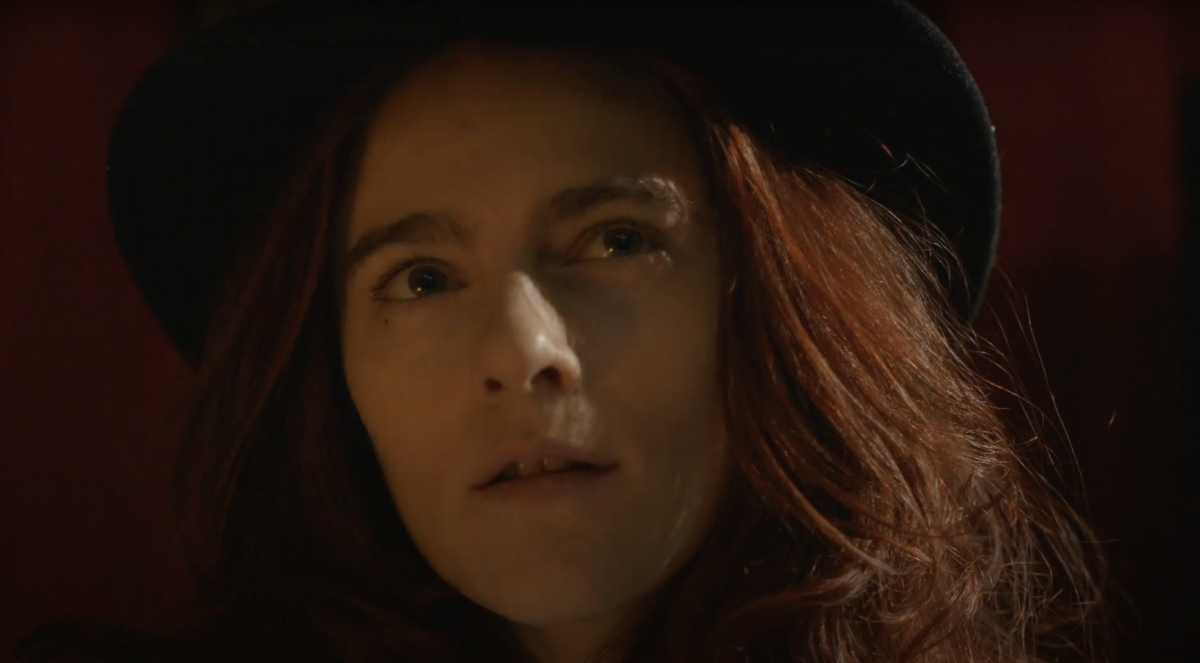 Close up of a woman with red hair and a black hat looking up, off camera.