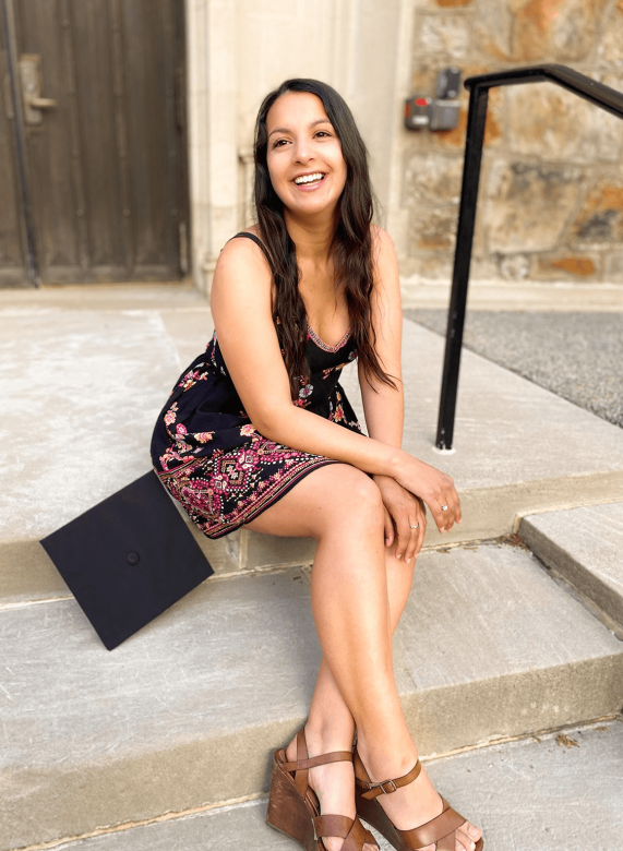 Anahí Alarcón sits on cement steps next to a graduate cap. Anahí wears a dress and is looking up, laughing. 