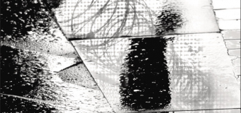Chrissy Kurpeski's book cover design for Shadows of Doubt: Stereotypes, Crime, and the Pursuit of Justice by Brendan O'Flaherty and Rajiv Sethi. The cover is black and white and looks like fractured glass covered in condensation. Behind the glass there is a shadowy figure walking to the right, away from a barbed wire fence.