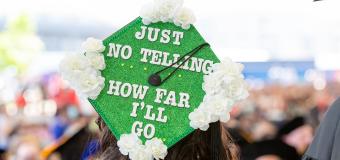 A graduate from the 2022 Commencement with a decorated cap. The cap is green with white flowers on its four corners and says "Just No Telling How Far I'll Go."