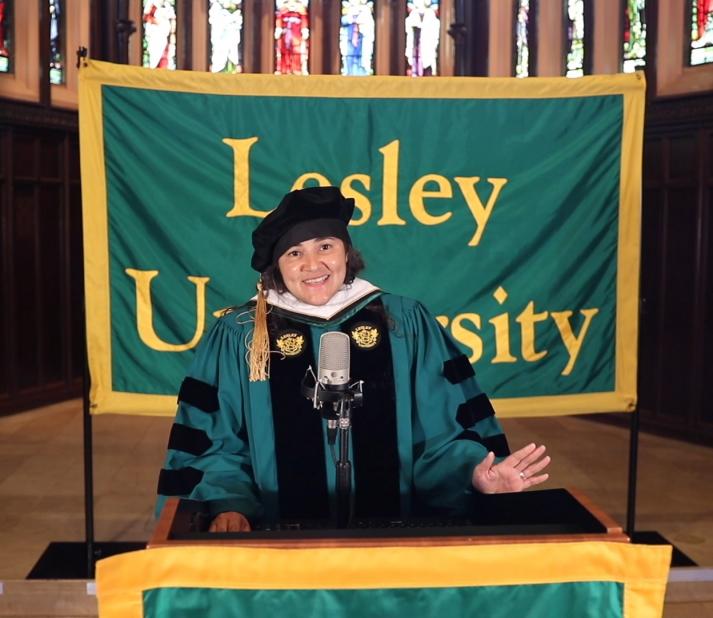A photo of Santos in her cap and gown for the 2021 virtual commencement ceremony. She is standing at a podium and there is a banner behind her that says "Lesley University."