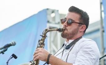 Man with sunglasses playing a saxophone on an outdoor stage.