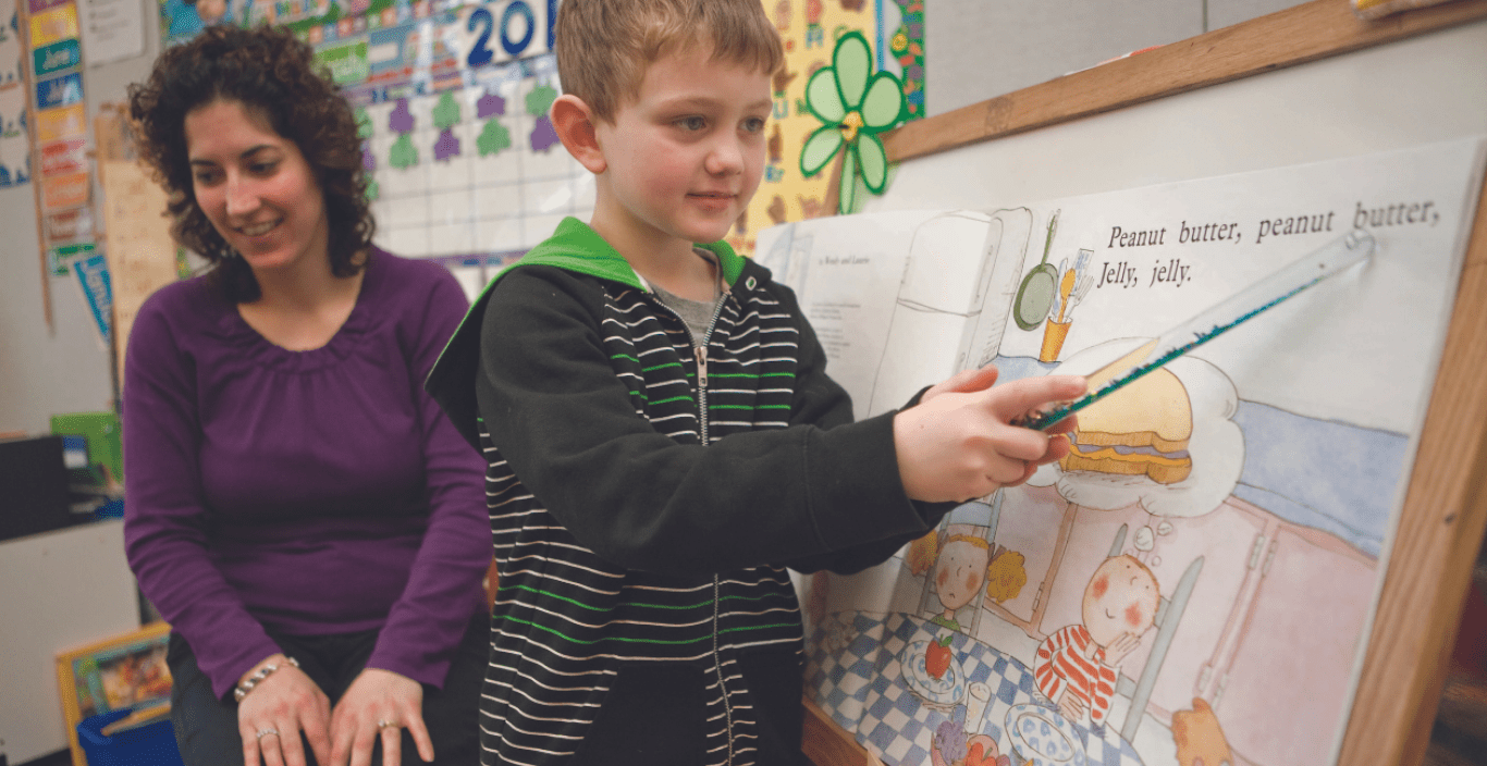 A photo with a kid in the foreground pointing to a board with a picture book image taped to it, with a teacher out of focus in the background. 