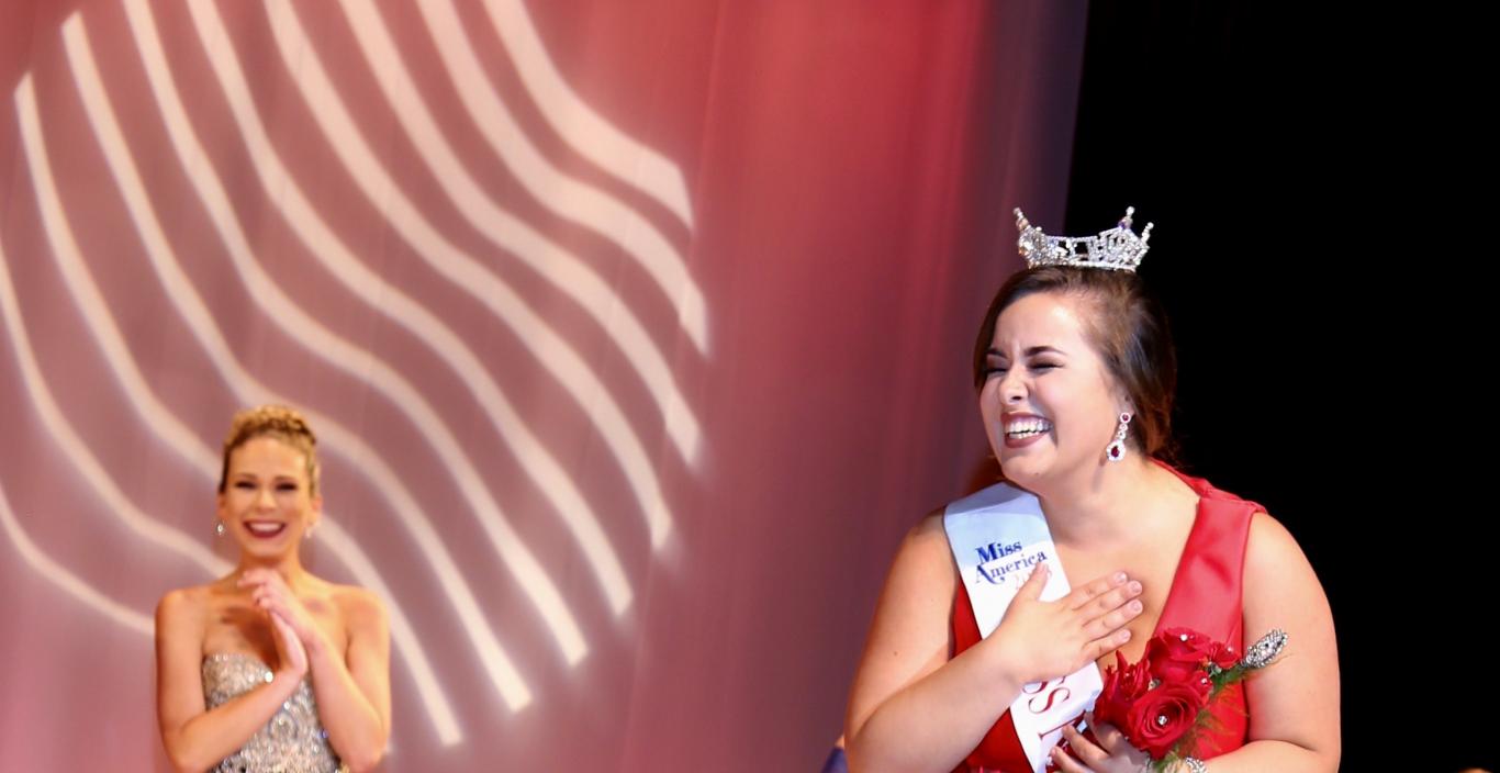 Sarah Achorn holds her hand to her heart in excitement as she wears the crown and Miss New Bedford sash on stage in her floor-length red dress.