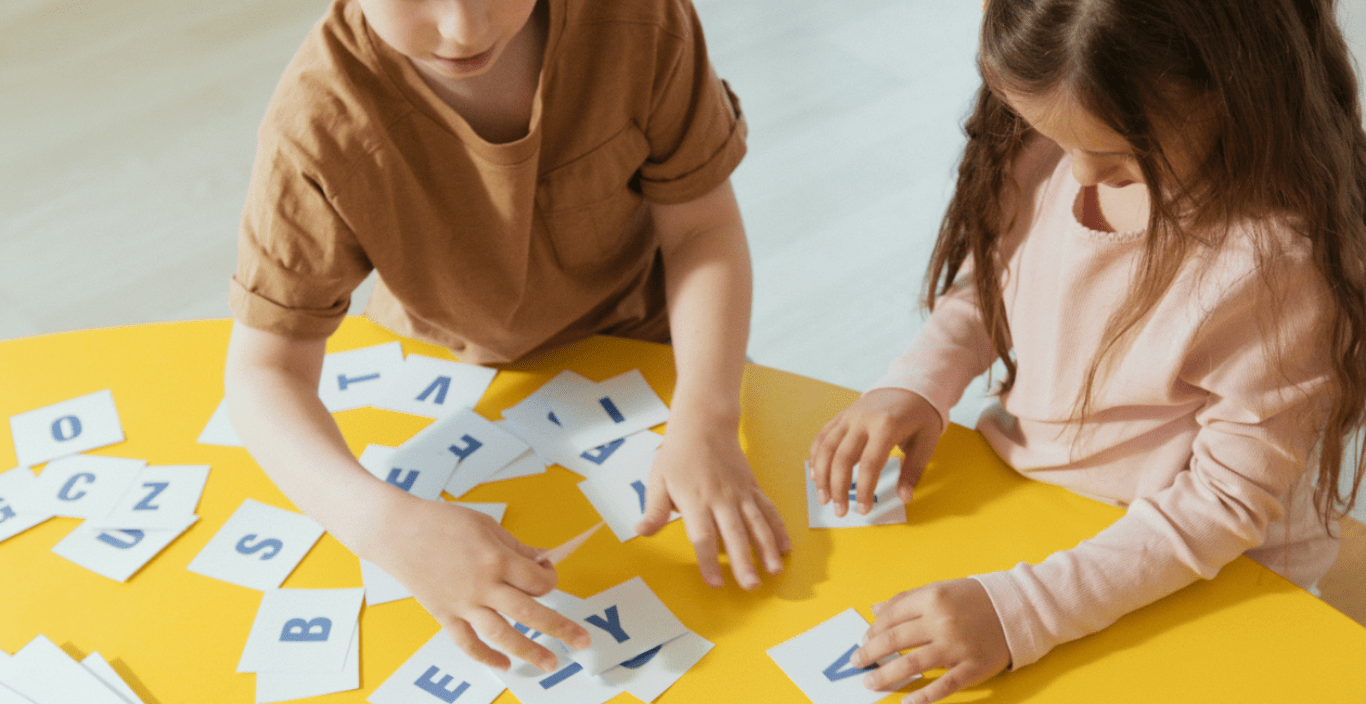 Two kids sitting at a yellow table with cut out letters on white papers strewn all over. The kids are assembling the letters to make words. 