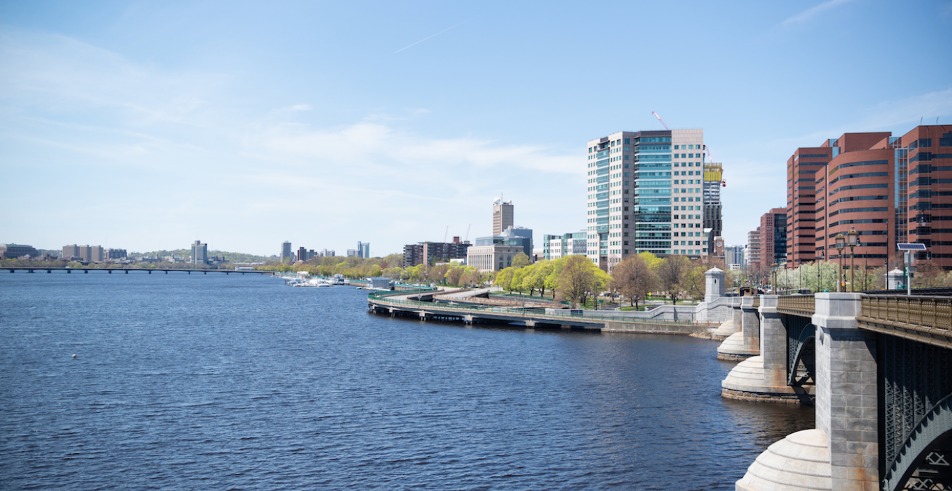 Photograph of the Charles River with blue sky and blue water and buildings of Cambridge in the background.