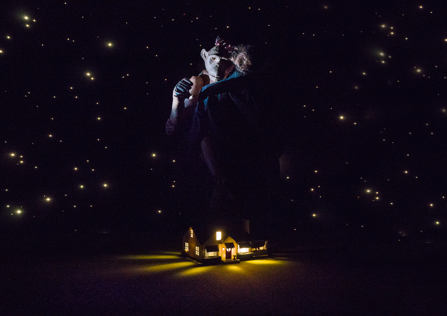 Image of chimpanzee puppet being held by a puppeteer with image of a house at night below them.