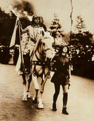 A sepia toned image of a woman dressed as Joan of Arc rides a horse lead by another woman.