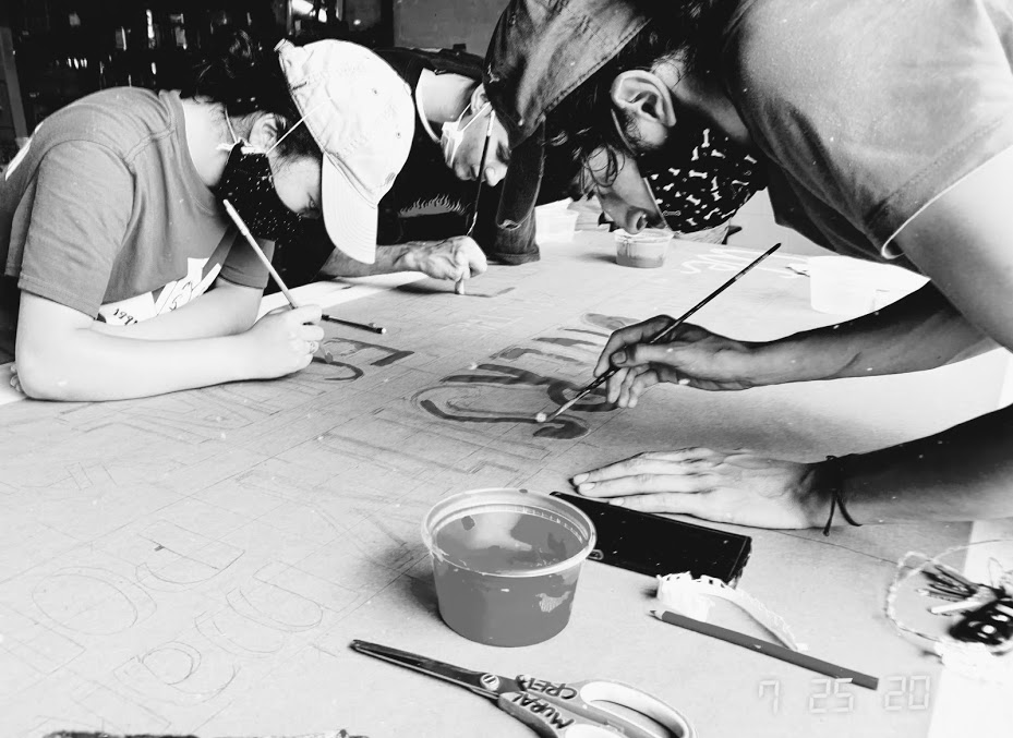 Black and white photo of masked people painting over a table.