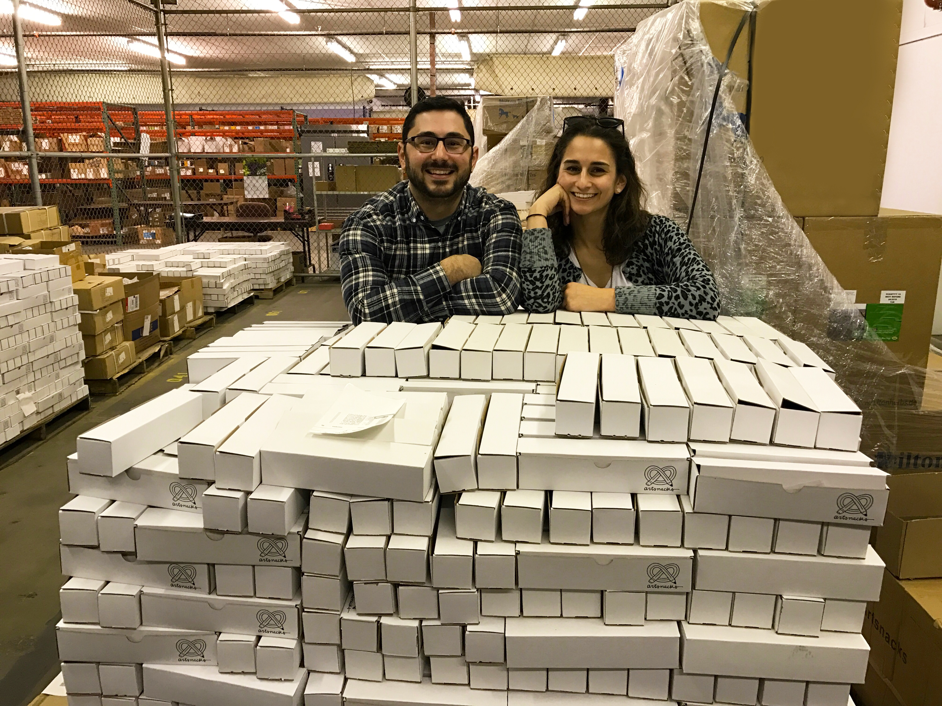 Sarah and Lee pose next to a large shipping pallet of ArtSnacks boxes. 