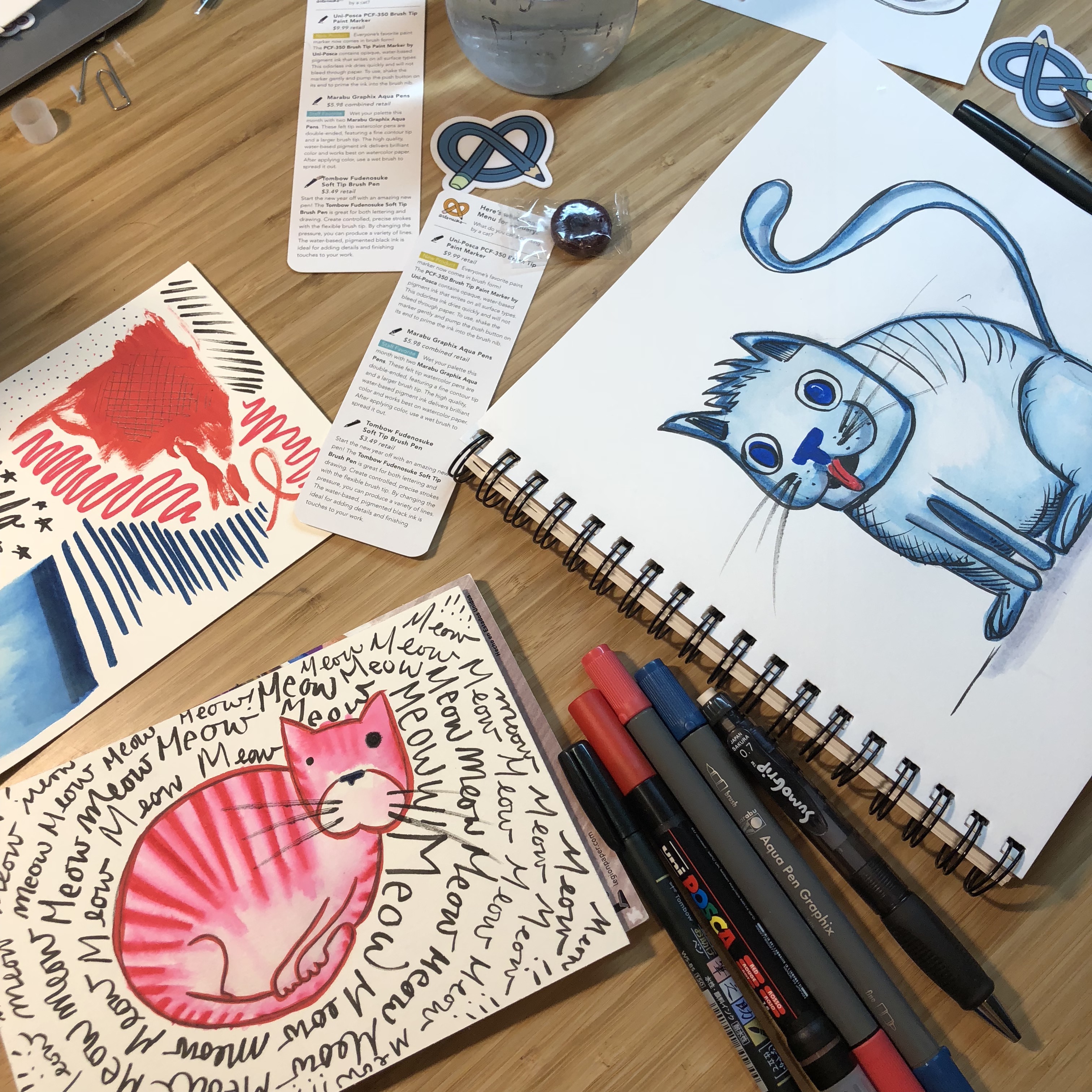 two pads of drawing paper with illustrated cats sit on a table surrounded by pens 