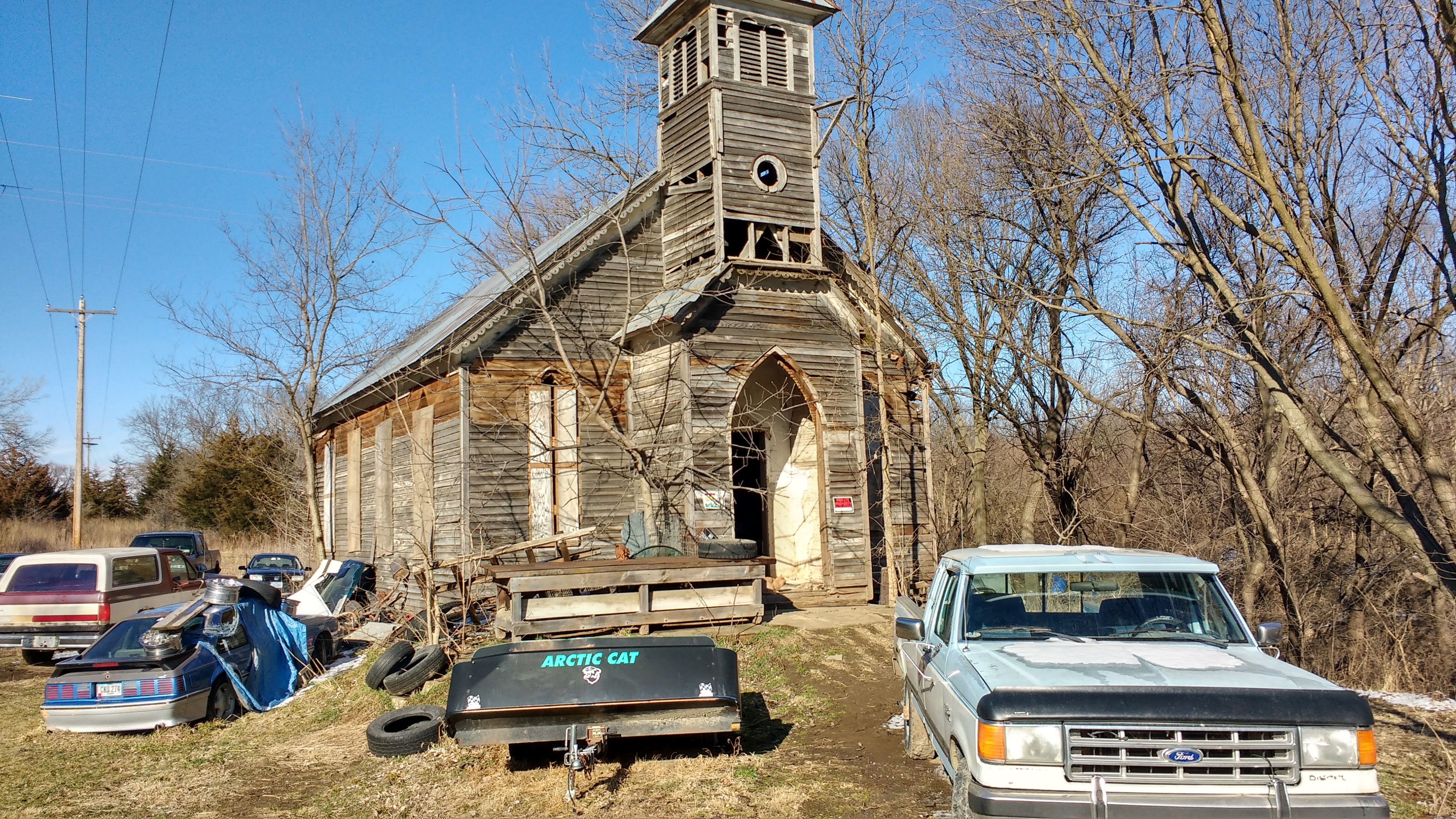 An old, abandoned church with broken down vehicles in front.