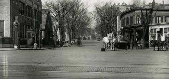 Old black and white photo of Harvard Square. Horses pull wagons in front of shops.