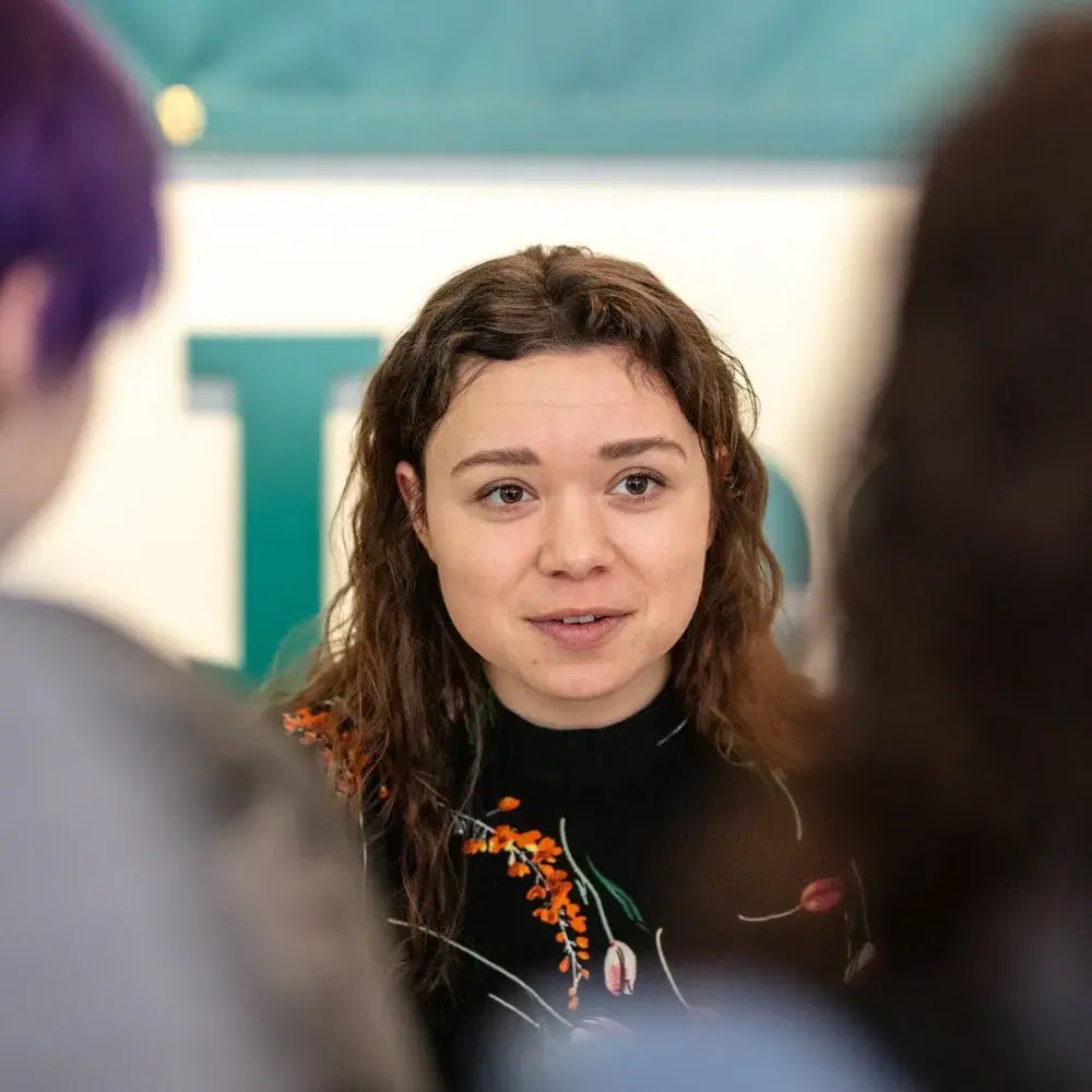 A young woman with long, wavy brown hair looks directly at the camera with a slight smile. She is wearing a black top with an orange and green floral pattern around the collar. The shoulders and backs of two other people are blurred in the foreground, framing her. In the background, a teal and white sign or logo is visible.
