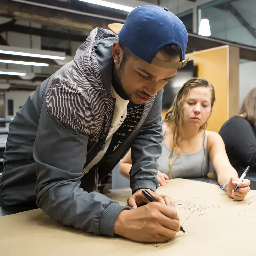 In a design studio, a group of four students gathers around a large table to collaborate on a drawing. A young man in a backwards cap leans over a large sheet of brown paper, actively sketching with a marker, while his three female classmates watch and contribute.