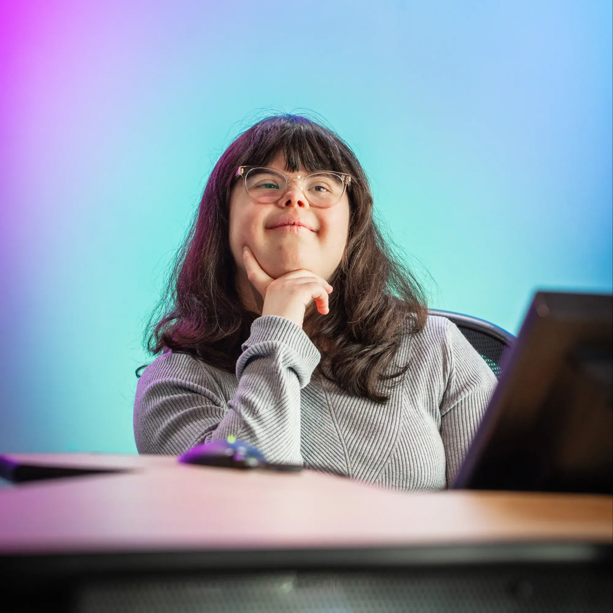 A young woman with long dark hair and glasses sits at a desk, resting her chin on her hand in a thoughtful pose. She is smiling slightly and wearing a ribbed light gray top. The background features a soft gradient of purples, blues, and teals, creating a vibrant and modern studio atmosphere. A computer monitor and mouse are visible in the foreground, suggesting a tech or media environment.