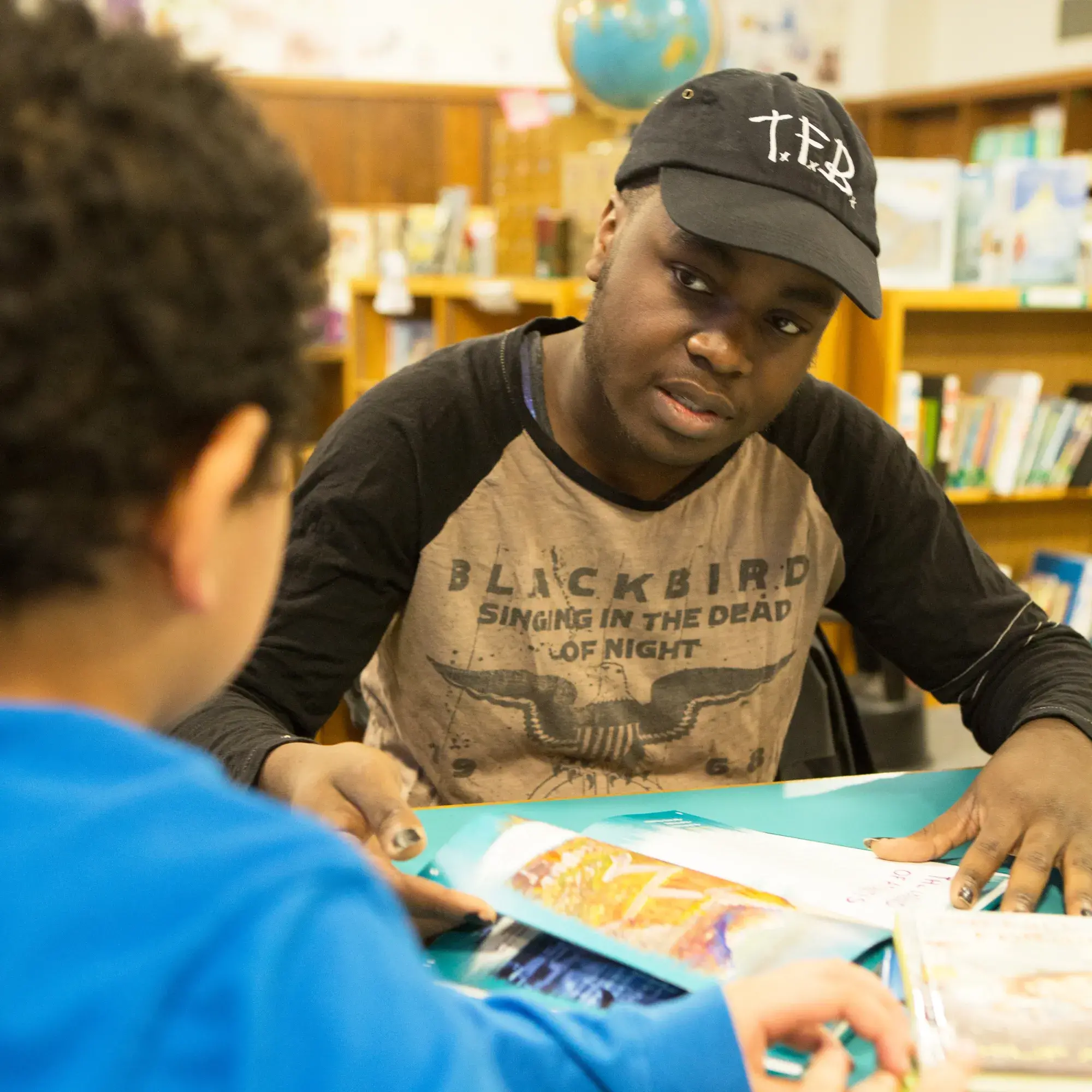 A person wearing a black cap and a shirt with the text "BLACKBIRD SINGING IN THE DEAD OF NIGHT" sits at a table in a library, interacting with a child in a blue shirt. They are looking at colorful, open books spread out on the table, with bookshelves and a globe visible in the background. The setting appears to be an educational or reading activity.