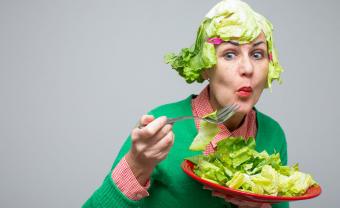 In this photo Eileen Powers in the right side of the image leaning forward looking left with her lips pursed chewing a piece of lettuce salad she is bringing towards her mouth with a fork. In the other hand she holds a red plate full of lettuce. She is wearing a red and white gingham collared shirt with a bright green cardigan over. She has her eyebrows raised and red lipstick on. Her wig is a bob made from large pieces of green lettuce with bangs and a hot pink hair clip pinned on each side.