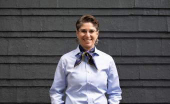 Jenn Steinfeld in a blue shirt and kerchief stands in front of a wall with wooden shingles