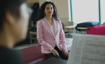 Woman in a pink blazer sitting on a bench in a classroom behind a woman playing the piano with sheet music. 