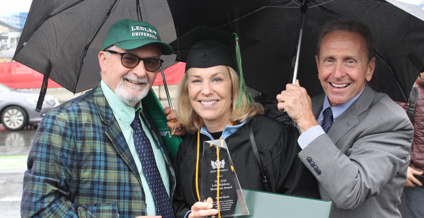 Anusia Hirsch wears her graduation robe and is stading with advisor Gene Ferraro and her husabnd under umbrellas holding her award and diploma