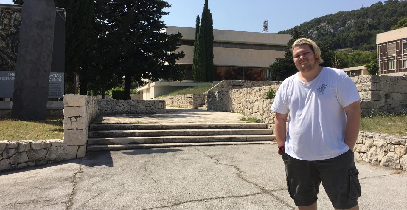 Tony Kecman in front of a museum in Croatia with stone buildings and green trees.