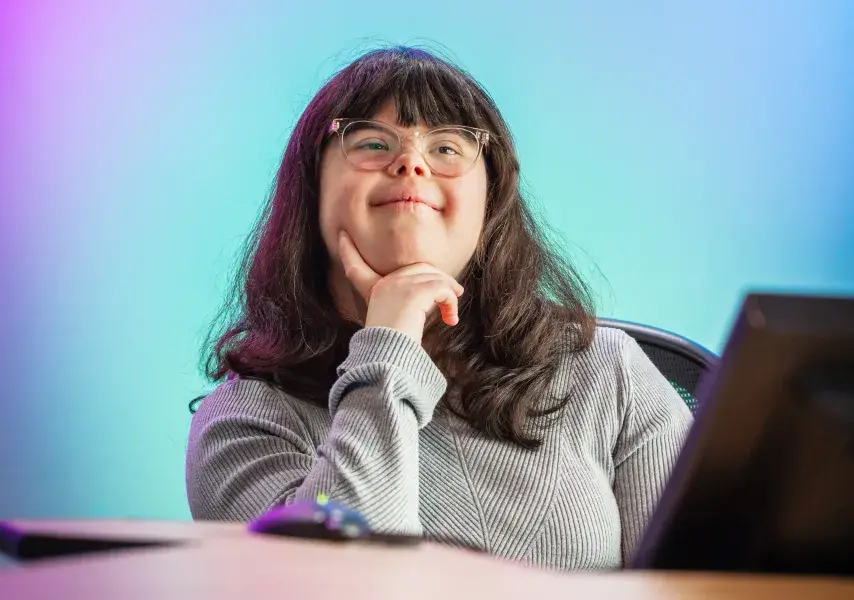 A young woman with long dark hair and glasses sits at a desk, resting her chin on her hand in a thoughtful pose. She is smiling slightly and wearing a ribbed light gray top. The background features a soft gradient of purples, blues, and teals, creating a vibrant and modern studio atmosphere. A computer monitor and mouse are visible in the foreground, suggesting a tech or media environment.
