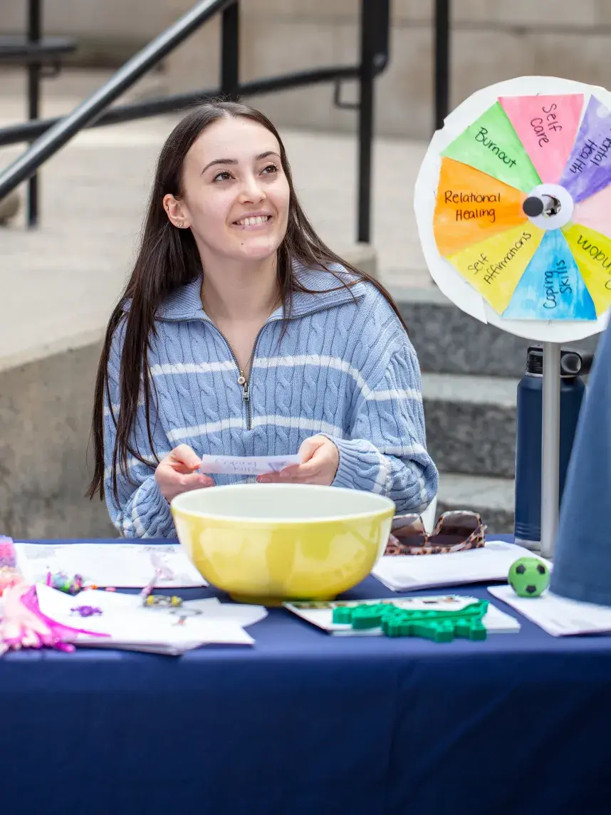 A friendly volunteer at a wellness fair engages with a visitor. She sits at an activity table featuring a colorful spinner wheel with positive themes and smiles as she holds a piece of paper, creating a welcoming and interactive atmosphere.